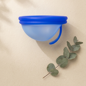 Blue menstrual cup on a beige background with eucalyptus leaves