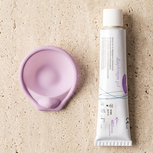 A white tube of daily moisturizer gel with purple accents is shown next to a lavender-colored silicone applicator pad on a textured beige stone background, with a sprig of lavender below.