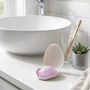 A pink and white clam-shaped soap dish holding a lavender soap bar, styled on a white bathroom counter next to a vessel sink, glass cup with a wooden toothbrush, and a small succulent.