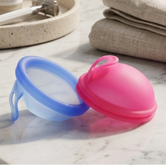 A pink silicone menstrual cup and a blue silicone cup lid resting on a white marble bathroom counter next to folded beige towels and skincare bottles.